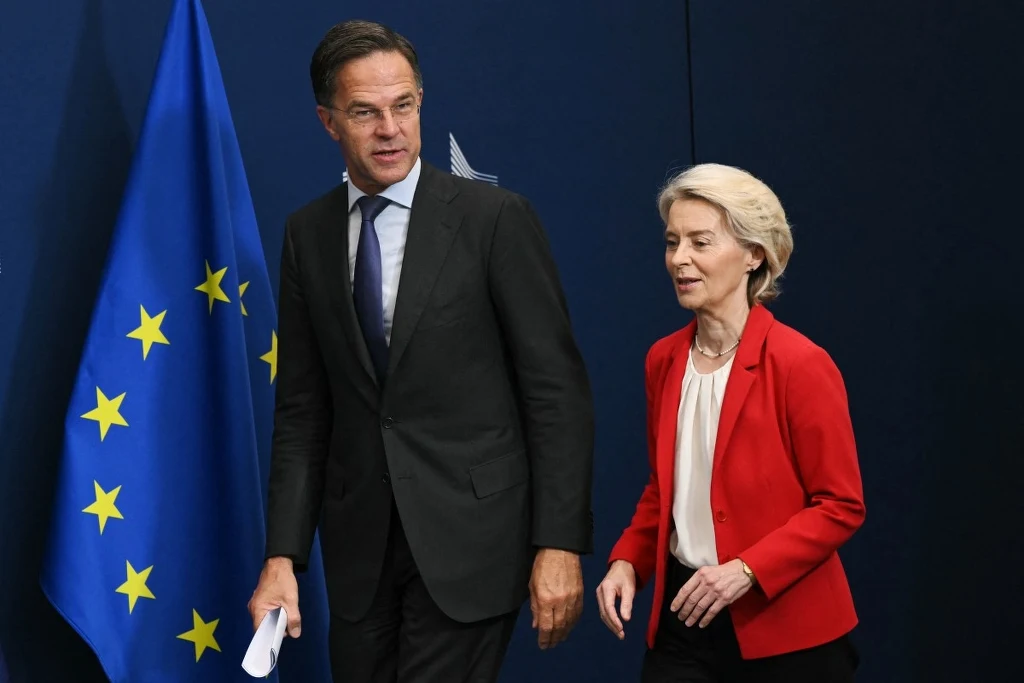 Sekretarz generalny NATO Mark Rutte i przewodnicząca Komisji Europejskiej Ursula von der Leyen Dwoje elegancko ubranych polityków przechodzi obok flagi Unii Europejskiej, mężczyzna trzyma kartkę papieru, kobieta ubrana w czerwoną marynarkę, ciemne tło podkreśla oficjalny charakter sceny.
