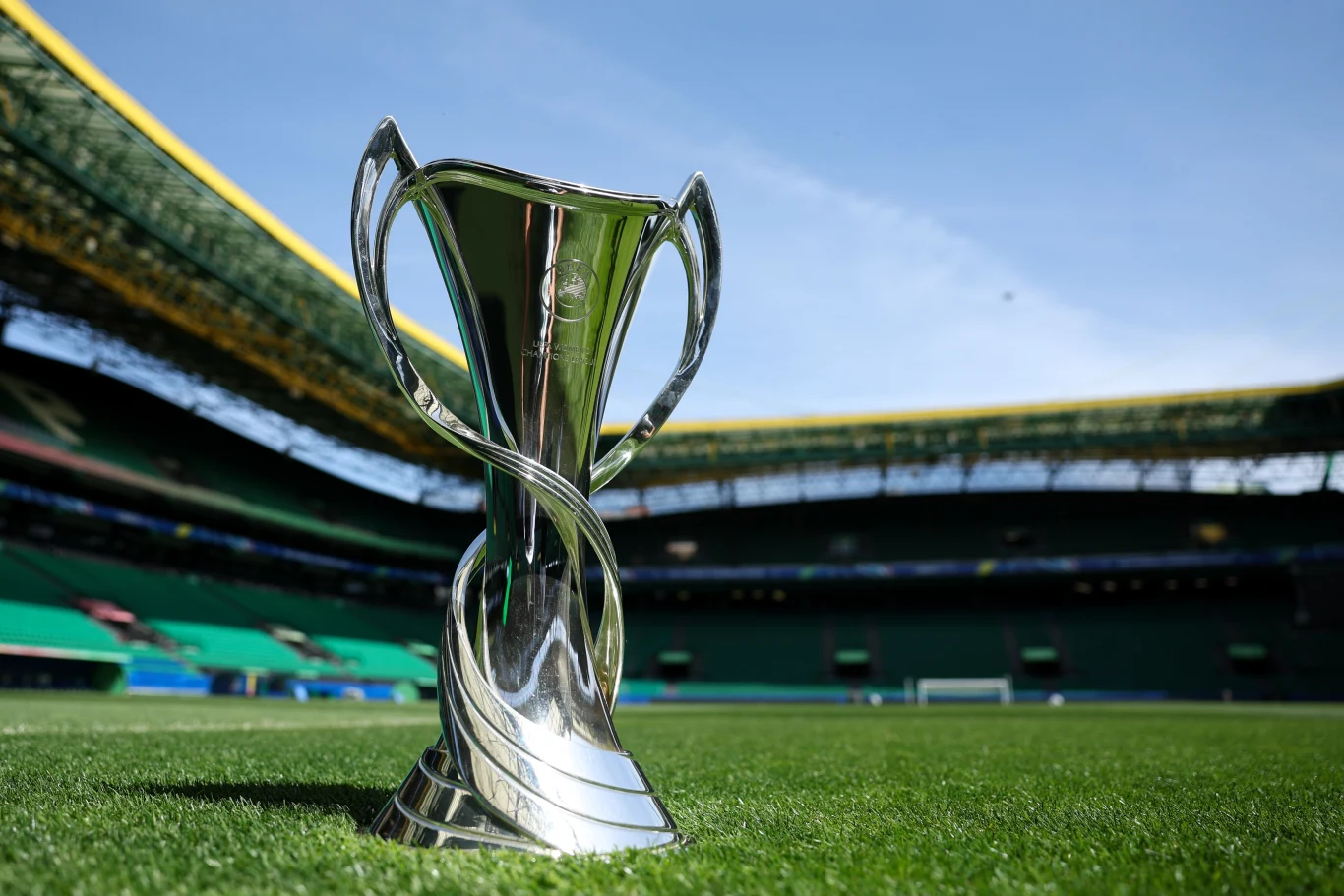 Arsenal, Lyon, Barcelona, Bayern, Wolfsburg, Paris Saint-Germain, Chelsea, Real Madryt, Roma, Juventus, Manchester United i inne drużyny powalczą o tytuł, a wszystkie mecze aż po finał w Oslo będzie można obejrzeć na żywo w Disney+. Srebrny puchar sportowy umieszczony na zielonej murawie stadionu, w tle trybuny i jasne niebo, sugerujące atmosferę oczekiwania na ważne rozstrzygnięcie sportowe.