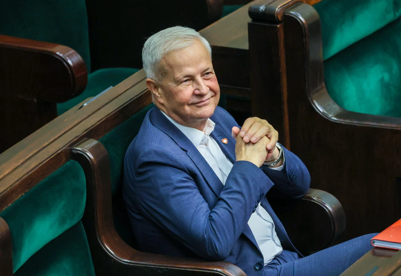 Starszy mężczyzna w niebieskim garniturze siedzi na sejmowej ławie, z lekkim uśmiechem na twarzy, złożone dłonie oparte o podbródek, zielone obicia ławek w tle.