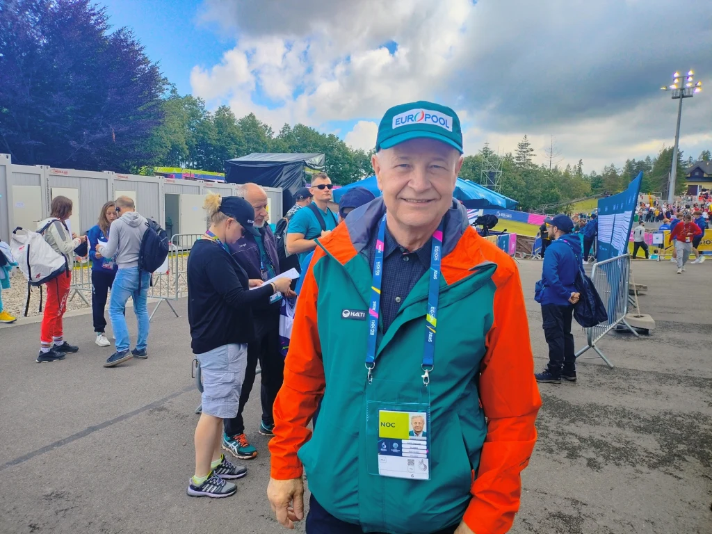 Apoloniusz Tajner Starszy mężczyzna w ubraniu sportowym i czapeczce z napisem Euro Pool stoi na tle imprezy plenerowej z licznymi uczestnikami, ogrodzeniem oraz banerami, w otoczeniu zieleni i nieba z chmurami.