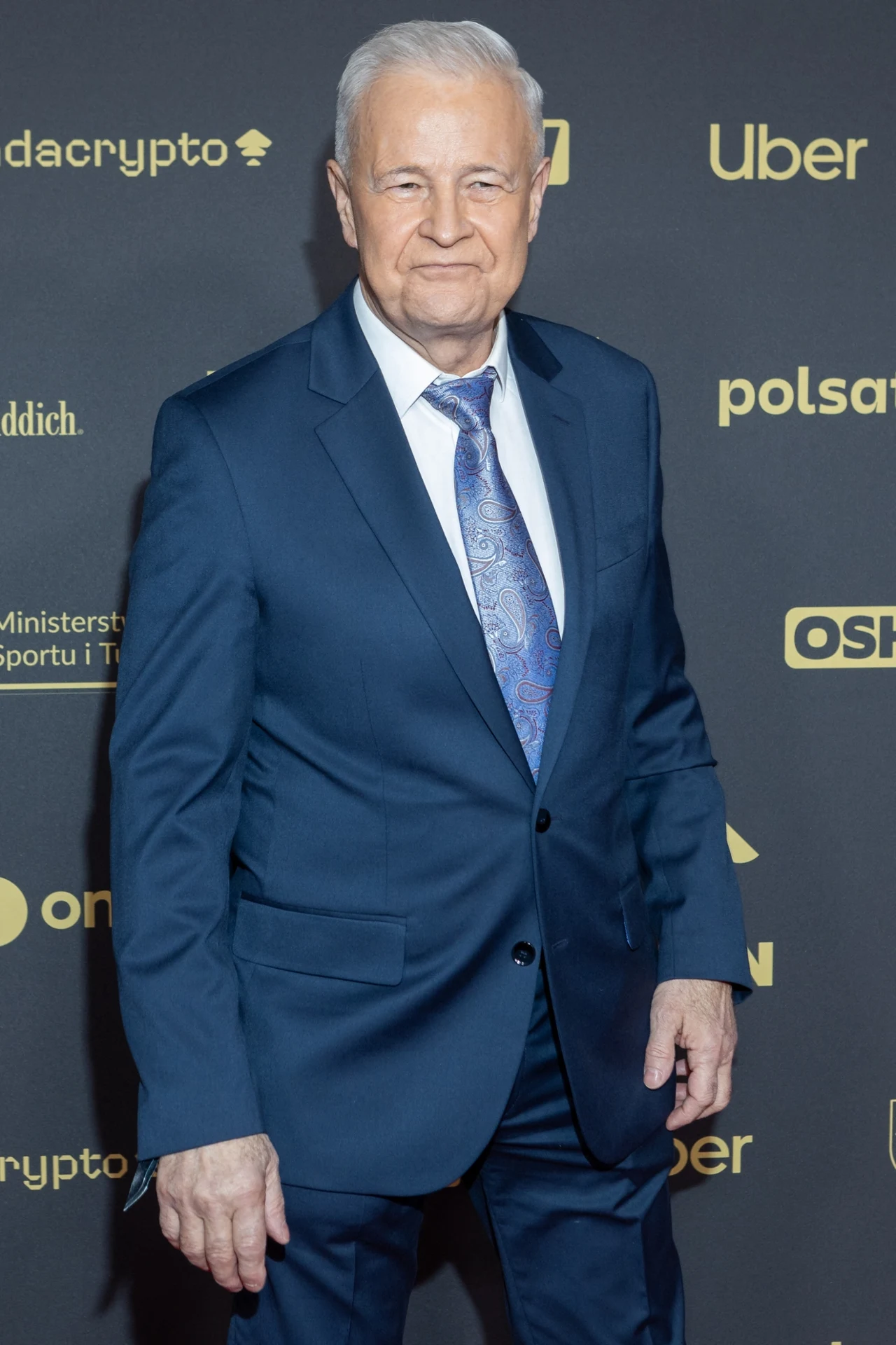 Elegancko ubrany mężczyzna w granatowym garniturze i błękitnym krawacie stoi na tle ścianki z logotypami różnych marek.