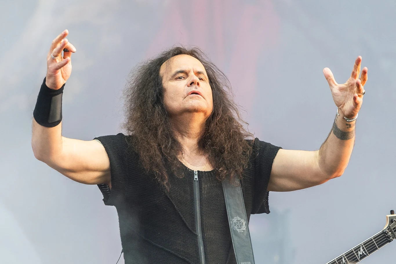 Na czele grupy Kreator stoi Miland "Mille" Petrozza Mężczyzna z długimi, ciemnymi włosami w czarnej koszulce wykonuje gesty dłońmi na scenie, widoczna część gitary, tło zamglone światłem koncertowym.