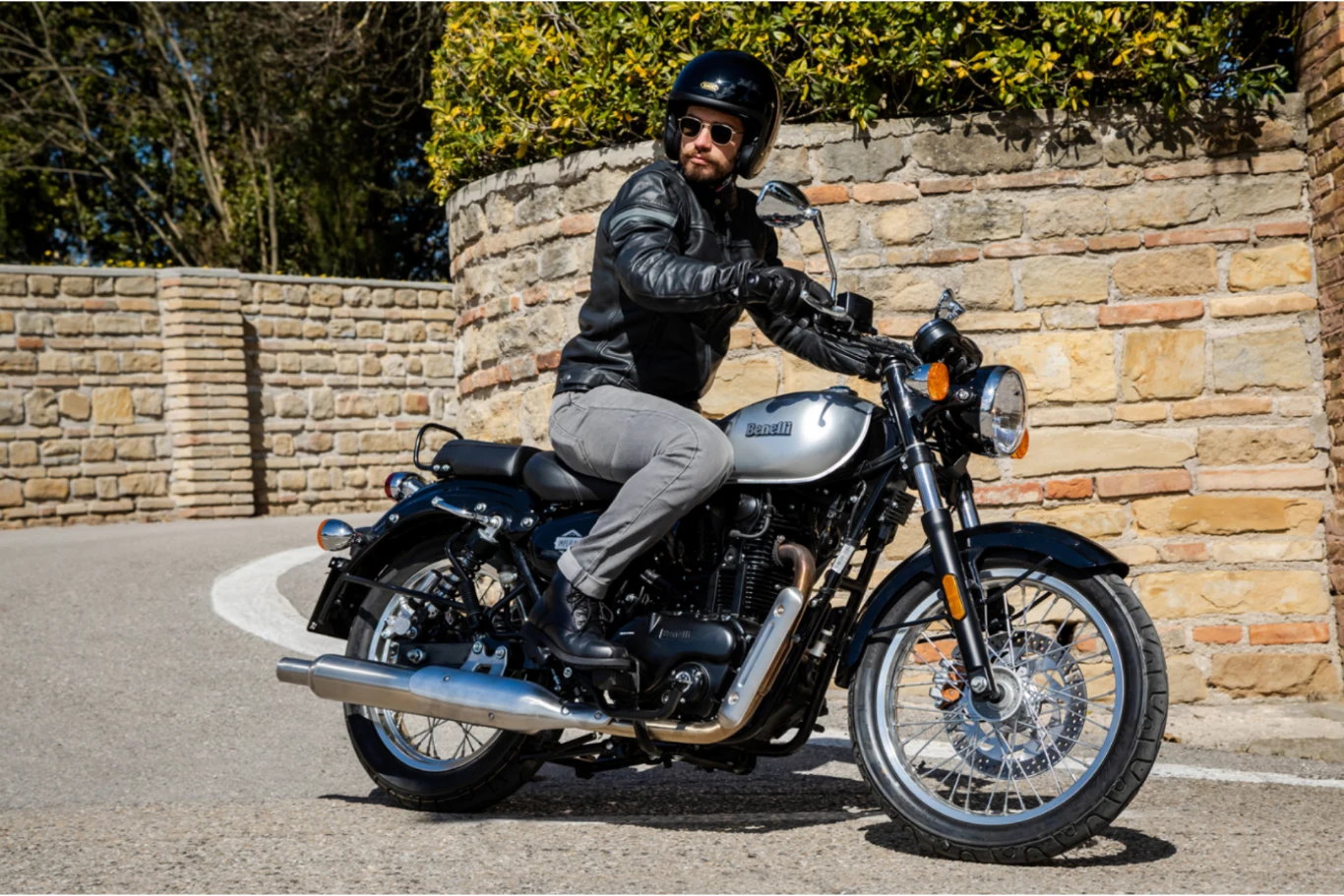 Ten motocykl nie może się nie podobać. Benelli Imperiale 400 wygląda ponadczasowo. Mężczyzna w czarnej kurtce i kasku jedzie srebrno-czarnym motocyklem po asfaltowej drodze przy kamiennym murze, w słoneczny dzień.