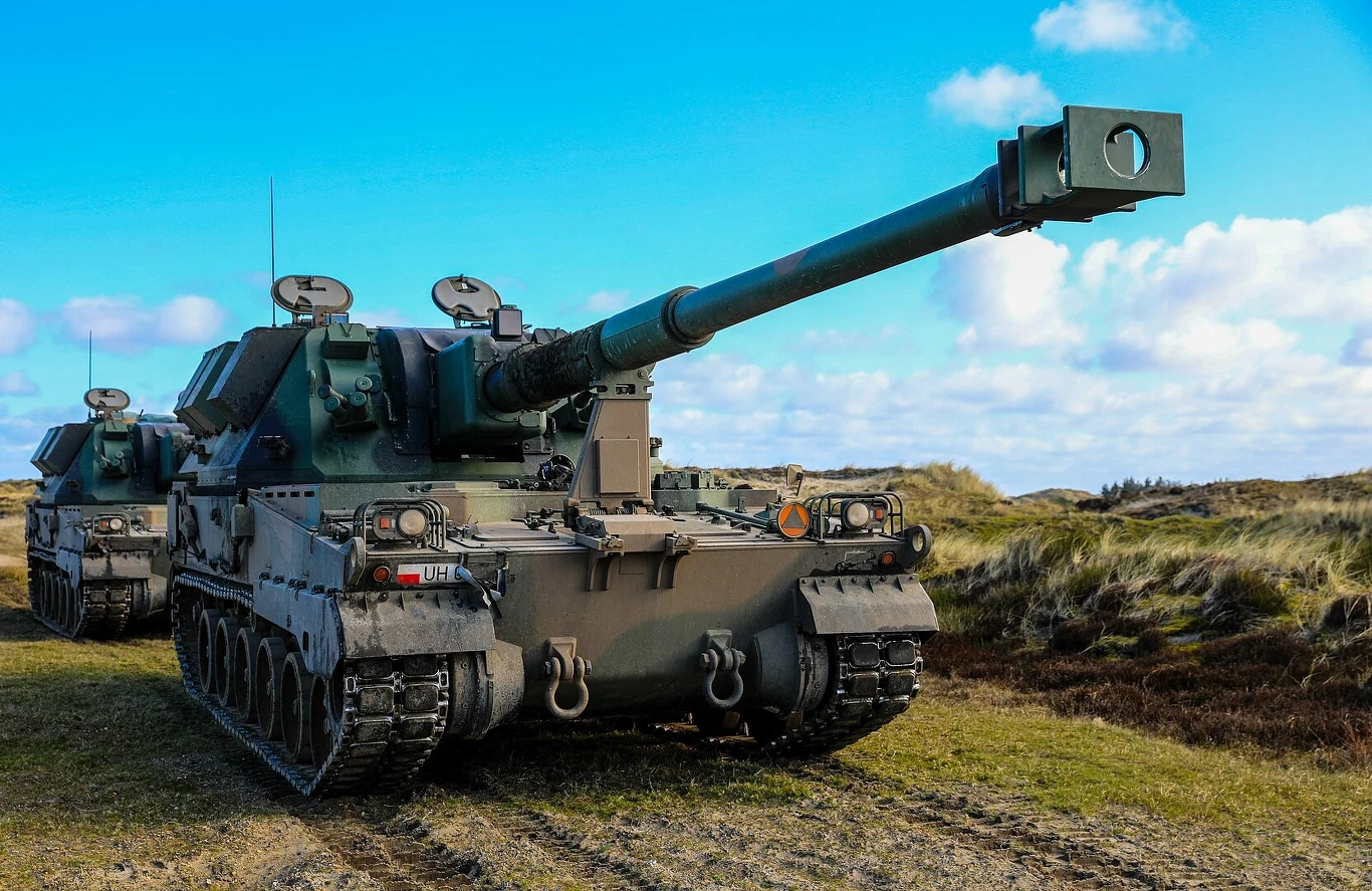 Armatohaubica Krab to artyleria samobieżna standardu NATO kalibru 155 mm. Wykorzystuje licencyjne podwozie koreańskich systemów K9 i wieżę z brytyjskiego AS90 Duża samobieżna armatohaubica wojskowa z wyraźnie widoczną lufą skierowaną do przodu, pojazd ustawiony na trawiastym terenie, nieopodal drugi taki sam pojazd, w tle pagórki i błękitne niebo z chmurami.