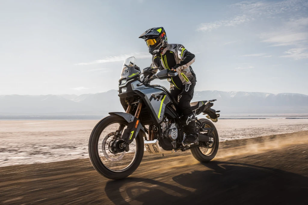 Motocyklista ubrany w profesjonalny strój oraz kask jedzie terenowym motocyklem po pustynnym bezdrożu, unosząc za sobą chmurę kurzu, w tle rozpościera się widok gór oraz otwartej przestrzeni.
