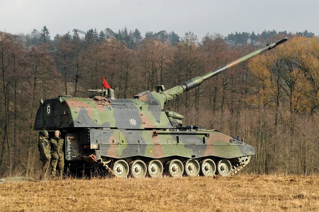 Samobieżna haubica w kamuflażu wojskowym ustawiona na otwartym polu z dwiema osobami w mundurach stojącymi obok pojazdu, w tle widoczne drzewa.