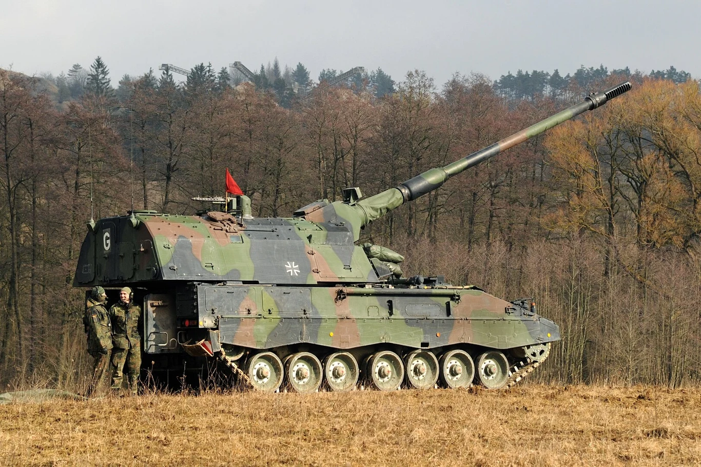 Panzerhaubitze 2000 to niemiecka armatohaubica, która zdobyła dużą popularność w Europie. Ich flotę posiada m.in. Grecja, Węgry, Holandia czy Chorwacja Samobieżna haubica w kamuflażu wojskowym ustawiona na otwartym polu z dwiema osobami w mundurach stojącymi obok pojazdu, w tle widoczne drzewa.