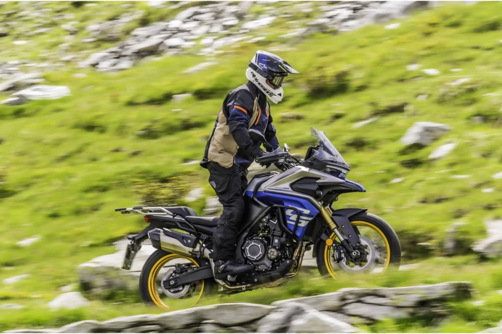 Motocyklista w pełnym stroju ochronnym jedzie niebiesko-żółtym motocyklem typu adventure po górzystym, zielonym terenie, otoczony kamieniami i trawą.