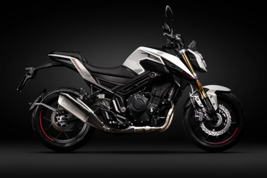 Sportowy motocykl typu naked z czarną ramą, białymi i srebrnymi elementami karoserii, masywnym tłumikiem oraz widoczną tarczową hamulcową z przodu, oświetlony na tle ciemnego tła.