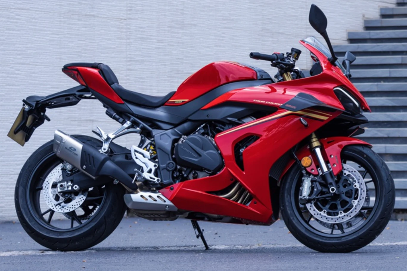 Motocykl w specyfikacji niemalże z WSBK może być w twoim garażu. SRK 800 RR kipi sportem. Czerwony motocykl sportowy zaparkowany obok schodów na tle jasnej ściany, z widocznymi nowoczesnymi elementami konstrukcji oraz aerodynamiczną sylwetką.