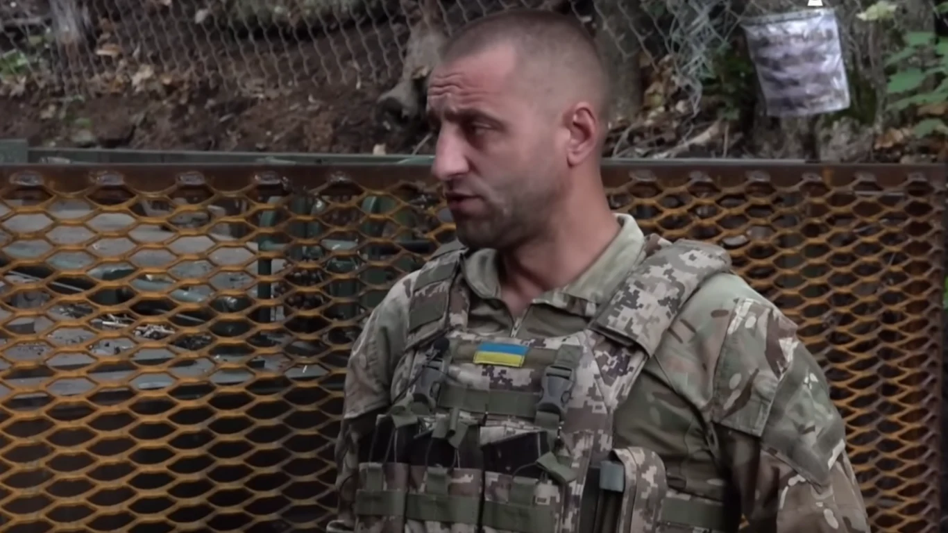 Serhij Mysiurla, dowódca ukraińskiej artylerii, w materiale Armia TV Mężczyzna w mundurze wojskowym z naszywką w barwach Ukrainy stoi na tle metalowej kratownicy, w tle widoczna siatka i fragmenty drzew.