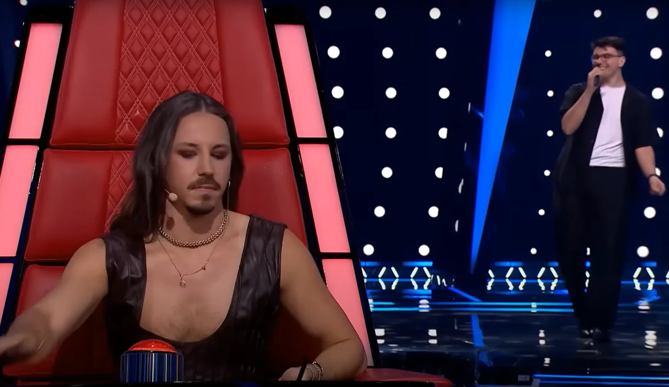 Stanisław Łukoński w "The Voice of Poland" zachwycił wszystkich trenerów Juror w charakterystycznym skórzanym stroju siedzi na czerwonym fotelu i naciska przycisk, w tle na scenie śpiewa mężczyzna ubrany na biało-czarno, scena oświetlona punktowymi światłami.