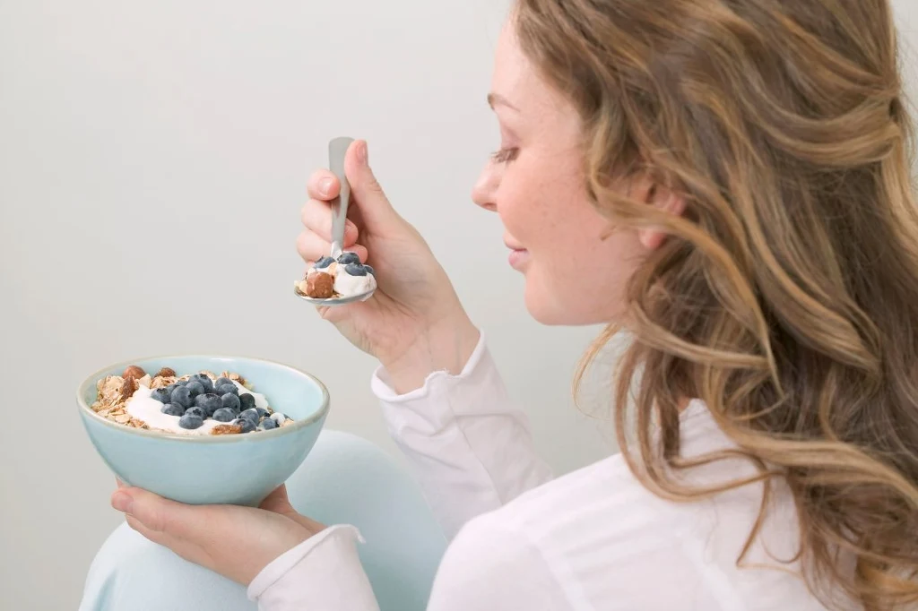 Nawet produkty, które uznajemy za zdrowe, mogą okazać się bezwartościowe i pełne szkodliwych substancji Młoda kobieta z jasnymi włosami trzyma w dłoni miskę jogurtu z borówkami i chrupkami, delikatnie uśmiechając się podczas nabierania łyżką porcji jedzenia.
