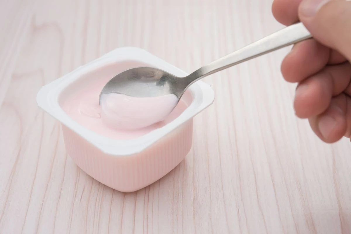 Różowy jogurt w plastikowym pojemniku oraz łyżka trzymana w dłoni, całość ustawiona na jasnym, drewnianym blacie.