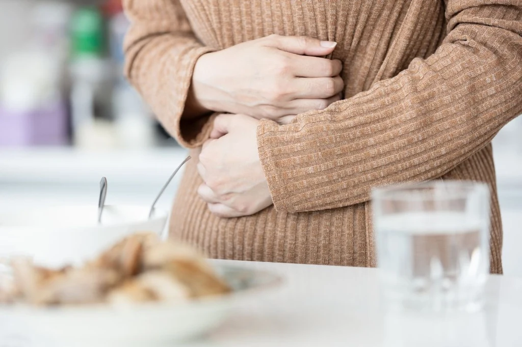 Osoba w brązowym swetrze trzyma ręce na brzuchu, siedząc przy stole, na którym stoi szklanka wody i talerz z jedzeniem.