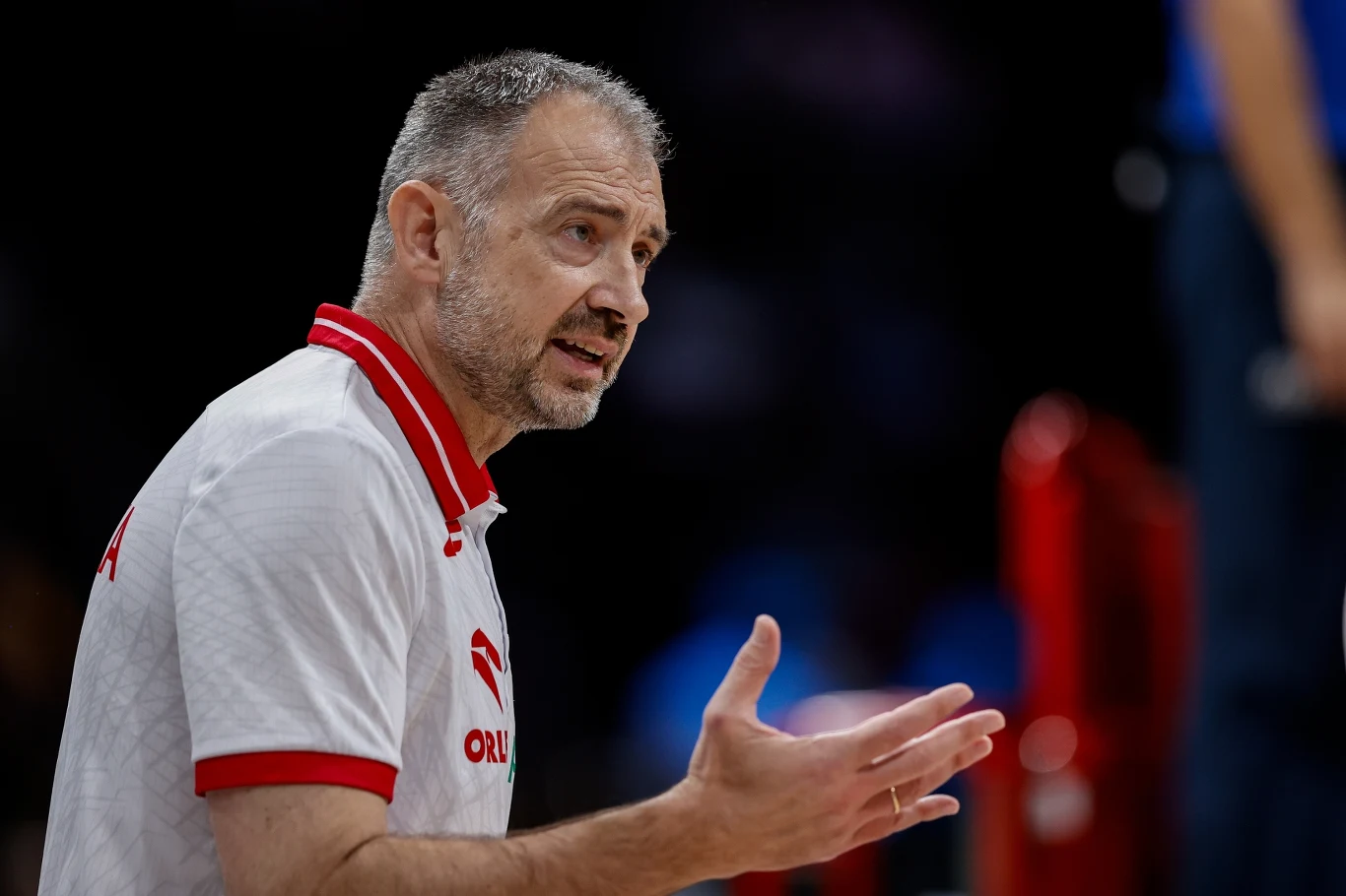 Nikola Grbić Trener w białej koszulce z czerwonymi akcentami gestykuluje, sprawiając wrażenie zaangażowania oraz komunikowania się podczas sportowego wydarzenia.