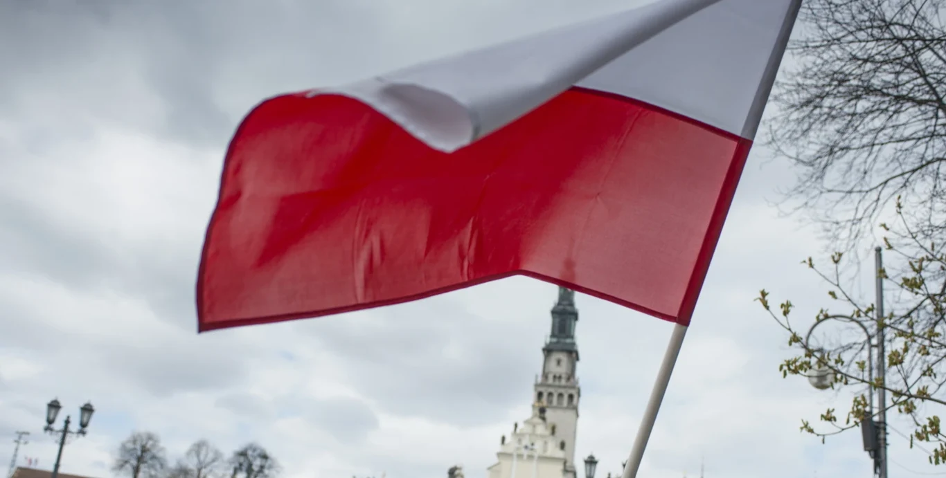 "kaajak" powalczy o finał na MŚ w Valorancie! Powiewająca flaga Polski na tle budynku klasztoru jasnogórskiego, pochmurne niebo, widoczna wieża z zegarem oraz gałęzie drzew