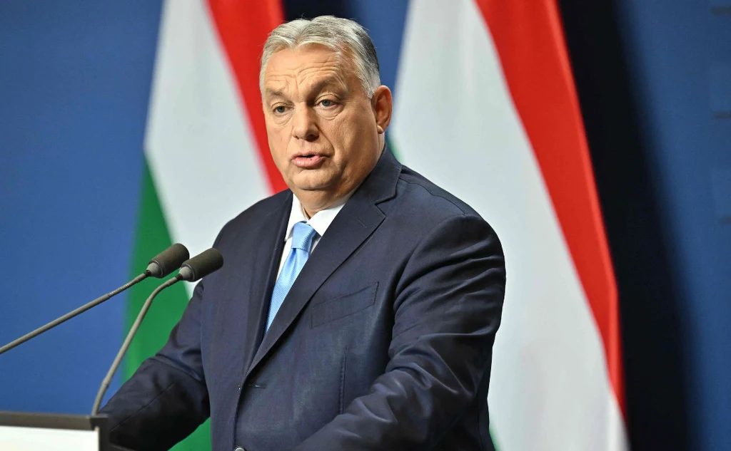 Premier Węgier Viktor Orban Mężczyzna w garniturze przemawia przy mównicy na tle biało-czerwono-zielonych flag, obok ustawione są mikrofony.