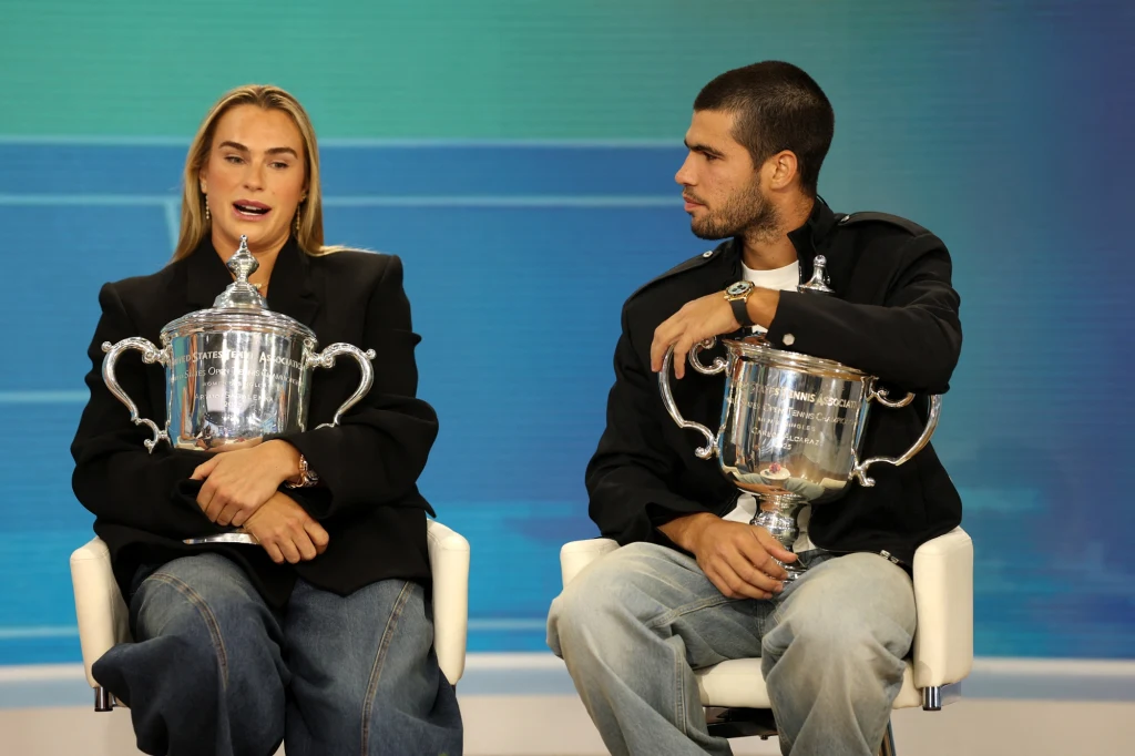 Aryna Sabalenka i Carlos Alcaraz wygrali US Open 2025, aktualnie przewodzą w rankingach WTA i ATP Dwie osoby siedzące na białych krzesłach, każda trzyma puchar, mają na sobie ciemne marynarki i dżinsy, tło utrzymane w niebiesko-zielonej tonacji.