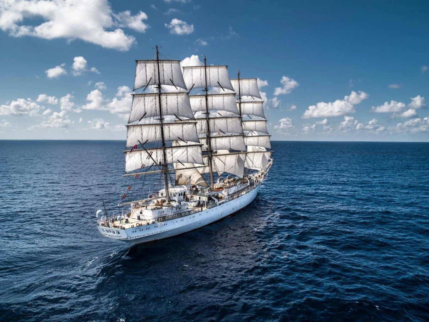 Duży żaglowiec z pełnymi żaglami płynący po spokojnym morzu, pod błękitnym niebem z pojedynczymi chmurami, otoczony bezkresną wodą.