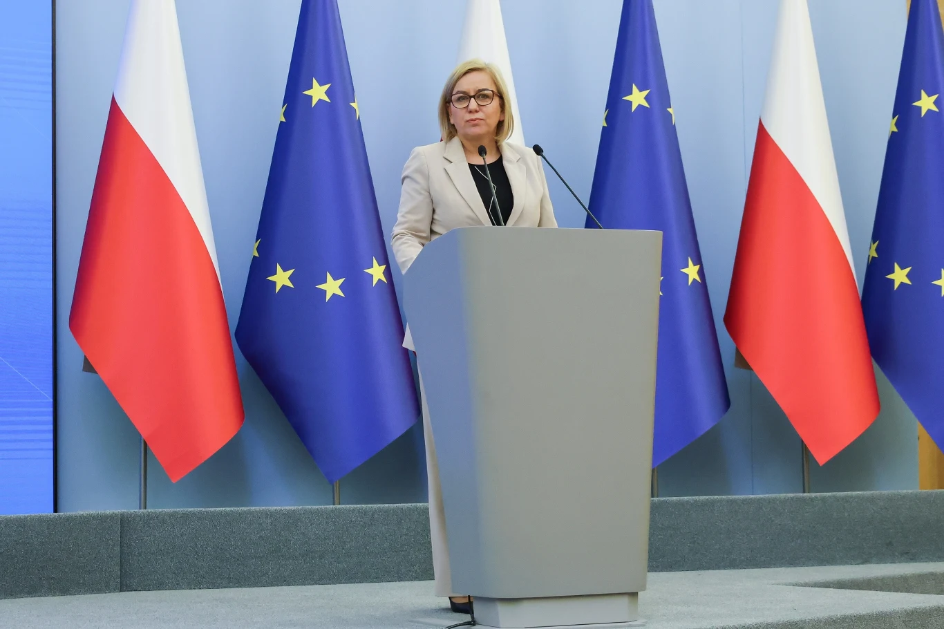System kaucyjny w Polsce rusza 1 października. Minister klimatu i środowiska Paulia Hennig-Kloska przybliżyła szczegóły jego działania Kobieta w jasnym garniturze stoi przy mównicy podczas oficjalnej konferencji prasowej, w tle widoczne flagi Polski oraz Unii Europejskiej.