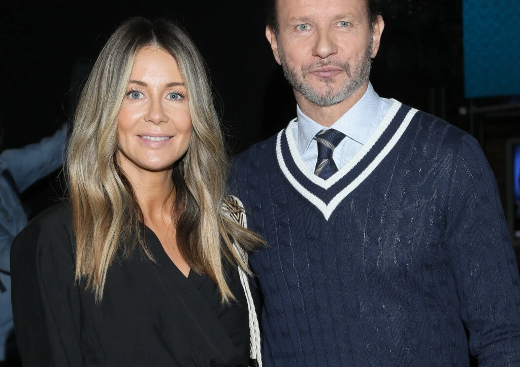 Małgorzata Rozenek-Majdan i Radosław Majdan Dwie uśmiechnięte osoby stojące obok siebie na tle ciemnego wnętrza, kobieta w czarnej kreacji z długimi blond włosami, mężczyzna w granatowym swetrze z białym kołnierzykiem i krawatem.