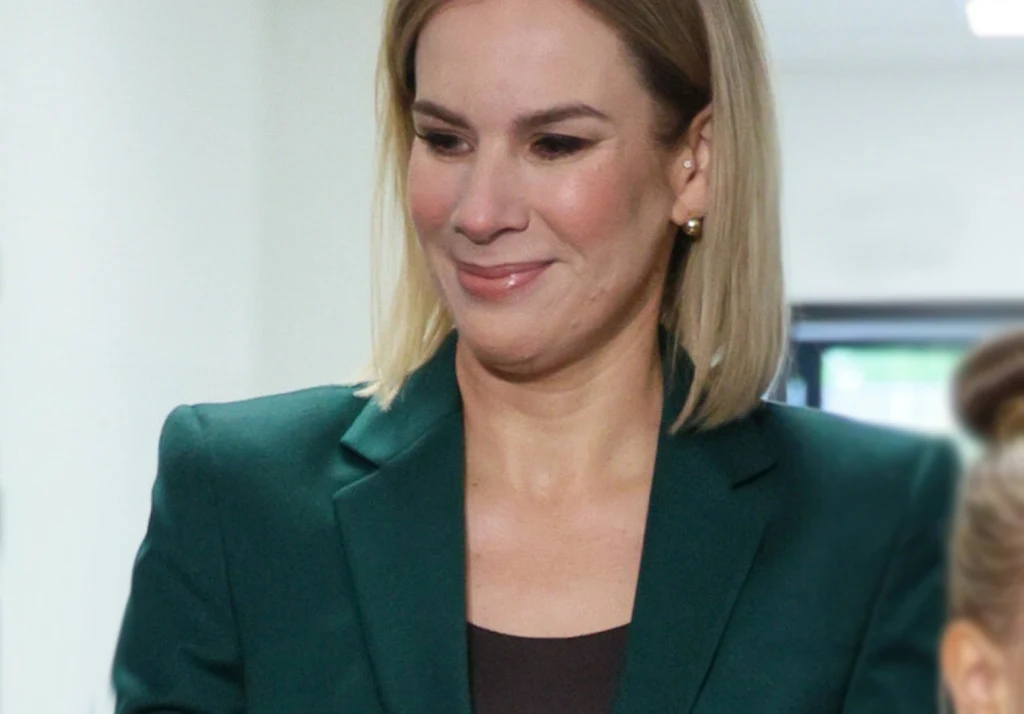 Kobieta w zielonej marynarce z jasnymi włosami, delikatnym makijażem, uśmiechem, stoi w jasnym wnętrzu.