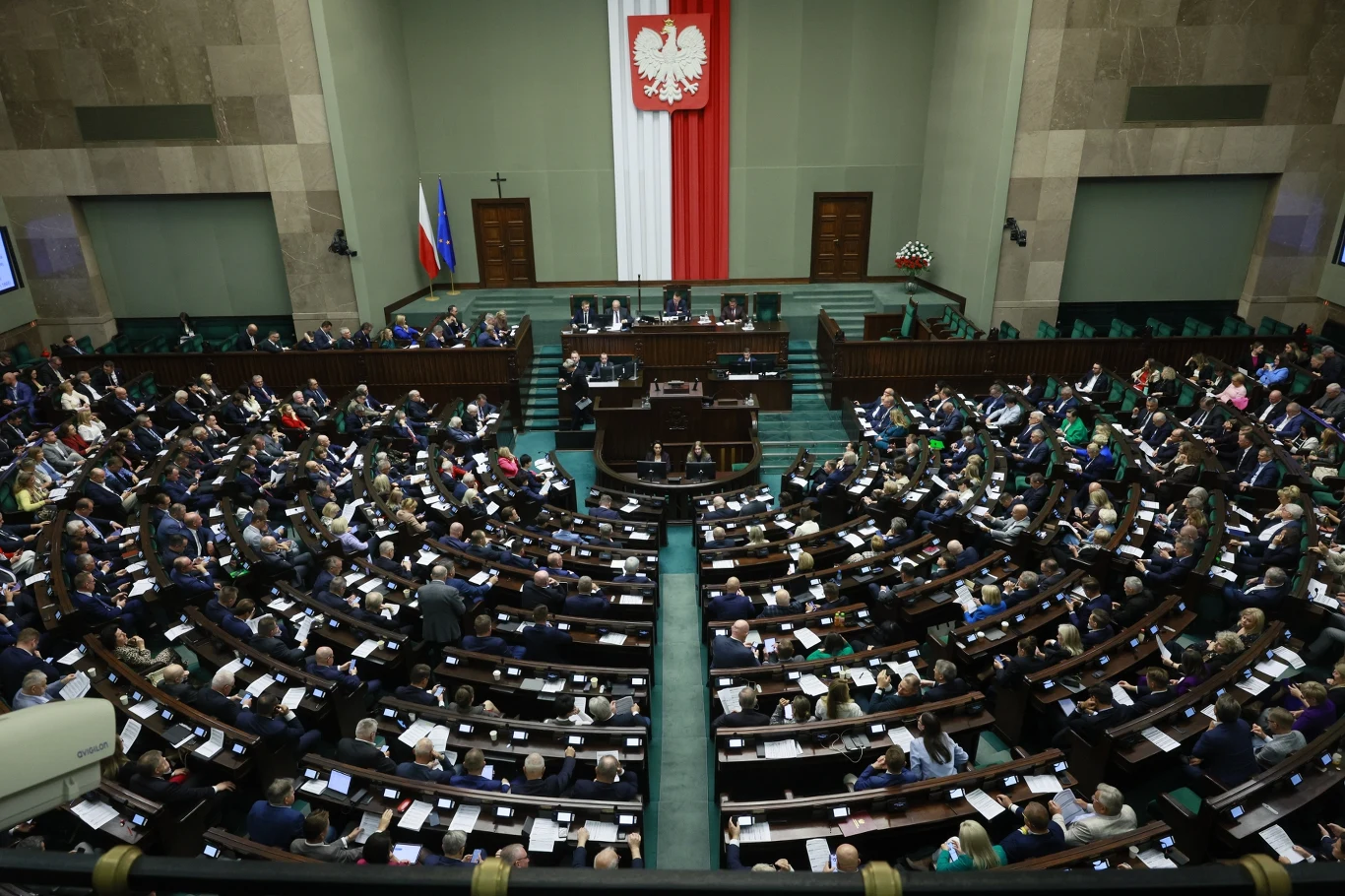 Sondaż. Kto wygrałby dzisiaj wybory parlamentarne? Sala obrad polskiego Sejmu zapełniona posłami, centralnie widoczna mównica z prezydium, w tle duża flaga Polski oraz godło państwowe, po bokach flagi Polski i Unii Europejskiej.