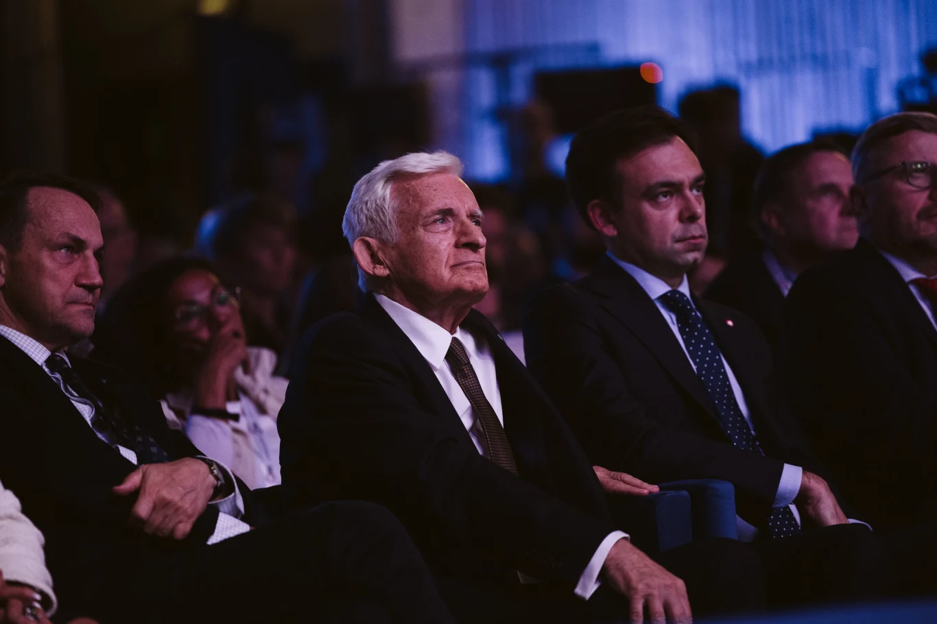 Grupa elegancko ubranych mężczyzn siedzących w rzędzie podczas formalnej konferencji, skupionych na obserwowaniu wydarzenia na scenie, w tle przyciemnione, nieostre światła.