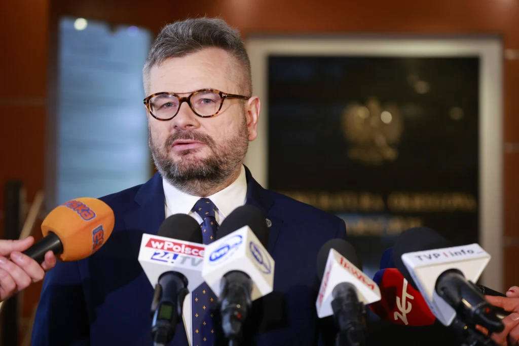 Mężczyzna w garniturze i okularach, stojący przed kilkoma mikrofonami z logotypami różnych stacji telewizyjnych podczas konferencji prasowej; w tle rozmyte godło i tablica urzędowa.