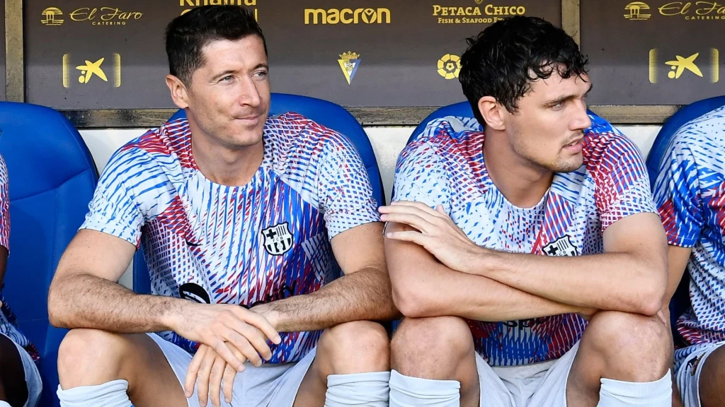 Robert Lewandowski i Andreas Christensen Dwóch piłkarzy siedzi na ławce rezerwowych w sportowych strojach, obaj w koszulkach z klubowym herbem, wokół nich widać fragmenty niebieskich siedzeń i tabliczki sponsorów stadionu.