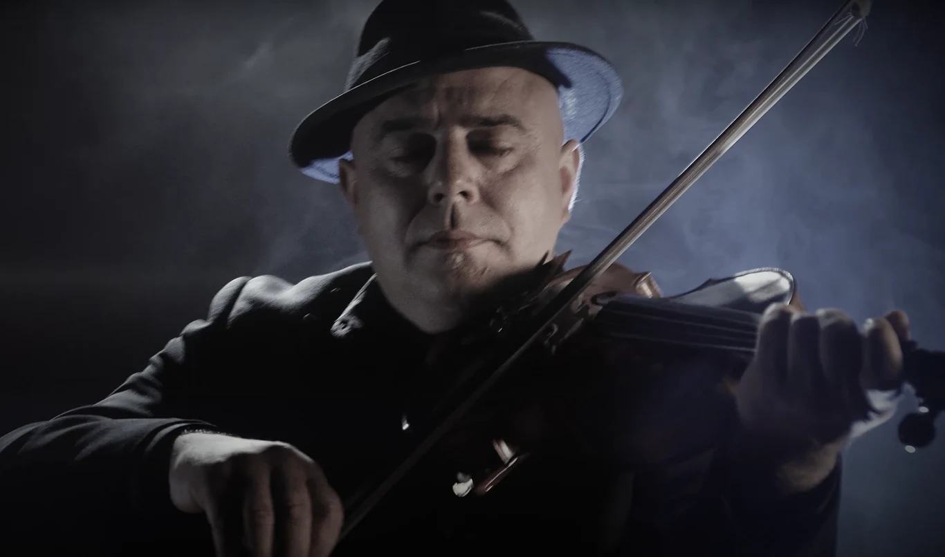 Andrzej Jarząbek "Fiś" jest liderem grupy FiśBanda Mężczyzna w ciemnym ubraniu i kapeluszu gra na skrzypcach, skupiony na muzyce, światło i dym budują nastrojowy klimat.