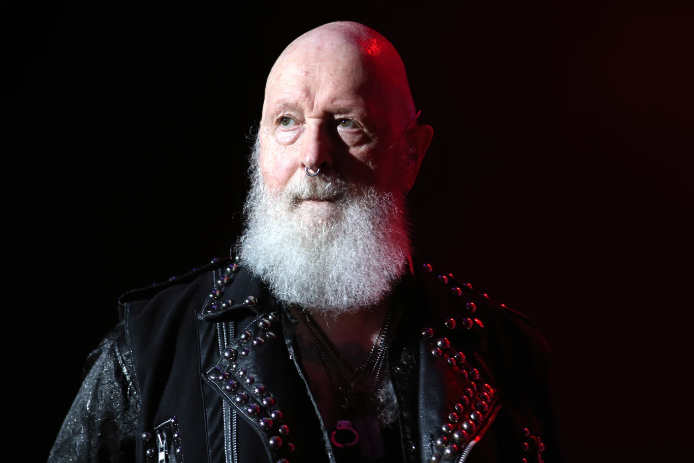 Rob Halford z Judas Priest Starszy mężczyzna z siwą brodą, ubrany w czarną skórzaną kurtkę z ćwiekami, patrzący przed siebie na tle ciemnego tła z czerwonym światłem.