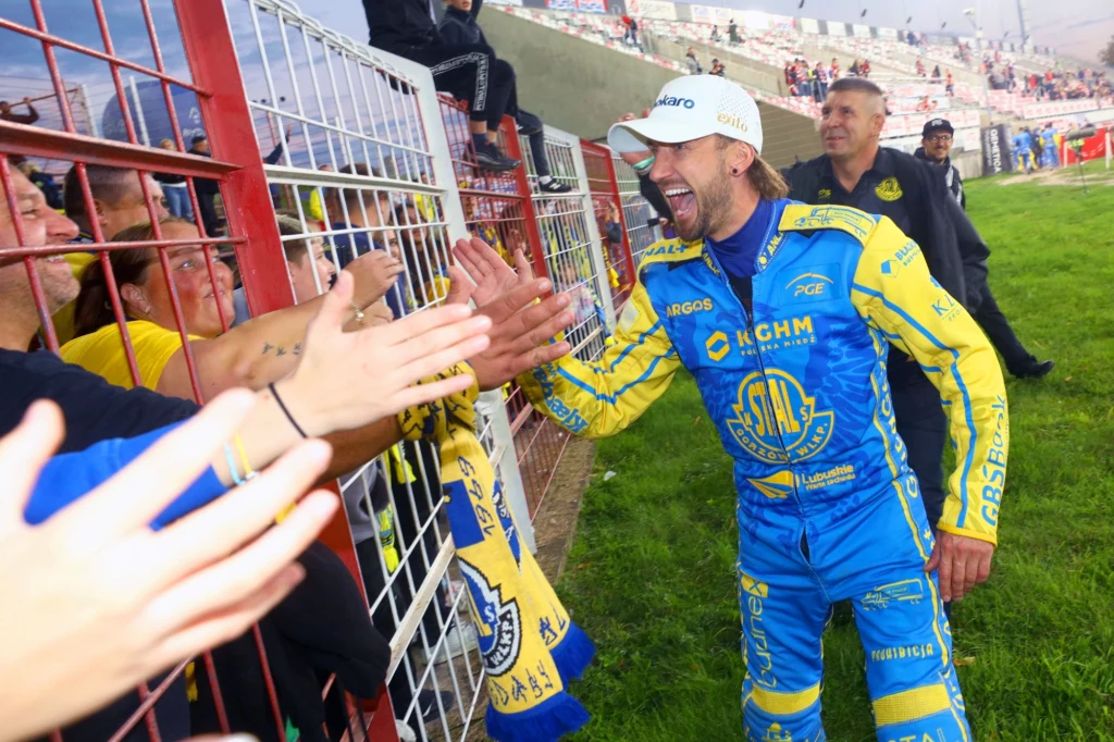 Anders Thomsen z kibicami po meczu w Bydgoszczy Zawodnik żużlowy w niebiesko-żółtym kombinezonie świętuje zwycięstwo z kibicami stojącymi za ogrodzeniem, przybijając piątki i okazując radość po zakończonym wyścigu.