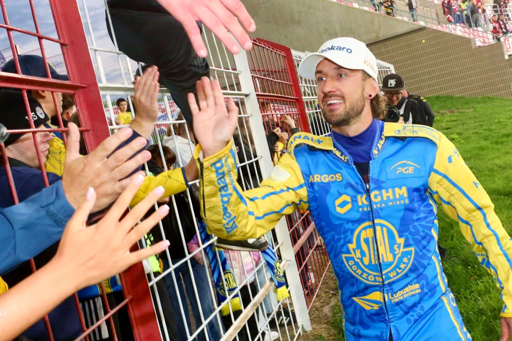 Uradowany Anders Thomsen zbija piątki z kibicami Zawodnik żużlowy ubrany w żółto-niebieski kombinezon oraz białą czapkę przybija piątki kibicom przez metalowe ogrodzenie na stadionie.