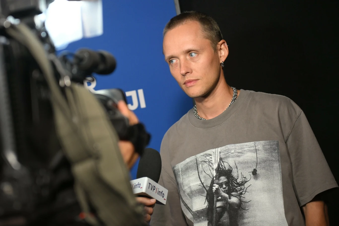 Bartosz Bielenia wcielił się w rolę lidera zespołu black metalowego Mężczyzna z krótkimi włosami noszący koszulkę z wyrazistym nadrukiem z grafiką twarzy, udzielający wywiadu przed telewizyjną kamerą oraz mikrofonem TVP Info, na tle niebieskiej ścianki.