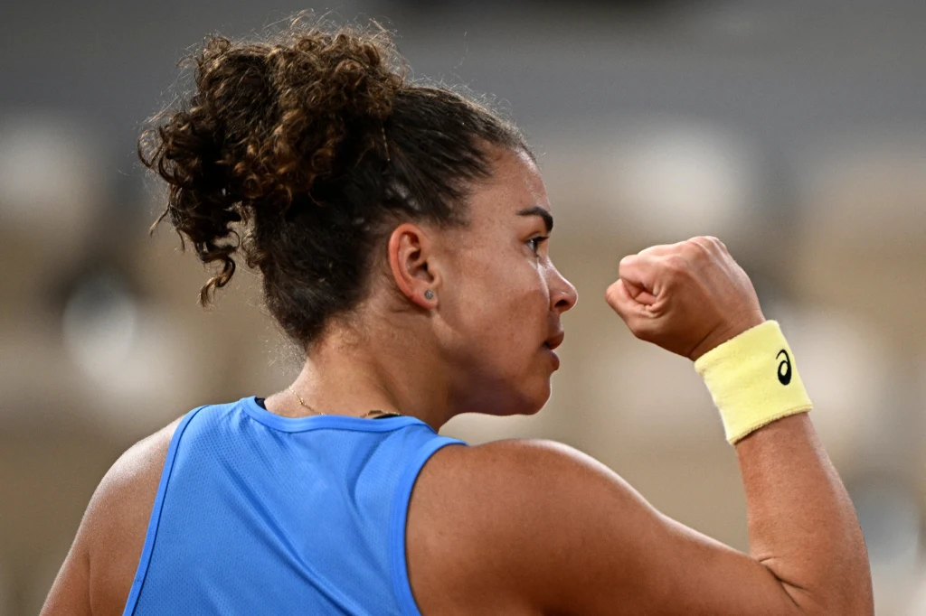 Jasmine Paolini Zawodniczka tenisa ubrana w niebieską koszulkę unosi zaciśniętą pięść w geście triumfu, wyrażając radość i determinację po udanym zagraniu. Jasna opaska na nadgarstku z widocznym logo marki.