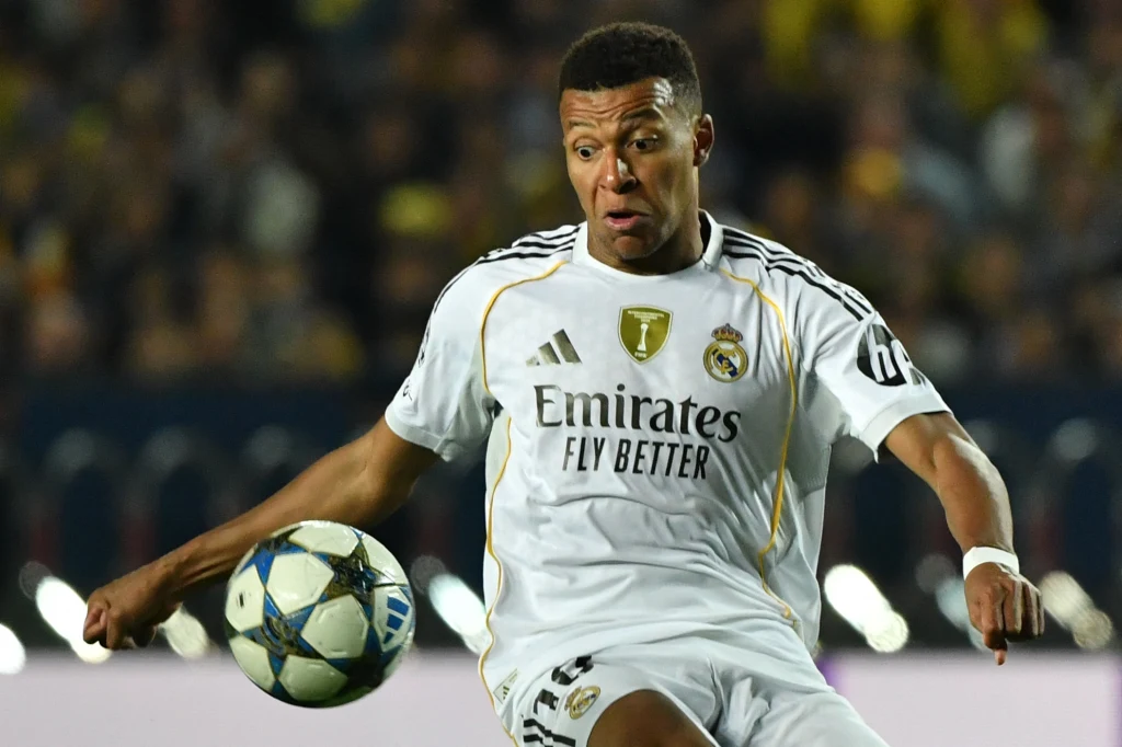 Kylian Mbappe w barwach Realu Madryt Piłkarz w białym stroju Realu Madryt koncentruje się na przyjęciu piłki podczas meczu na stadionie, w tle widoczna rozmyta publiczność.