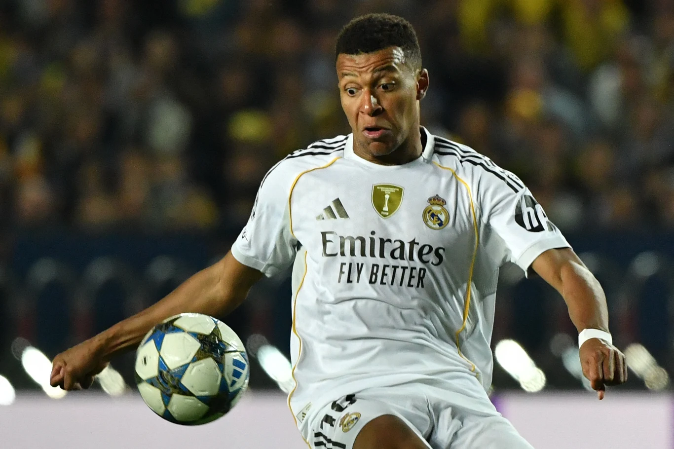 Piłkarz w białym stroju Realu Madryt koncentruje się na przyjęciu piłki podczas meczu na stadionie, w tle widoczna rozmyta publiczność.