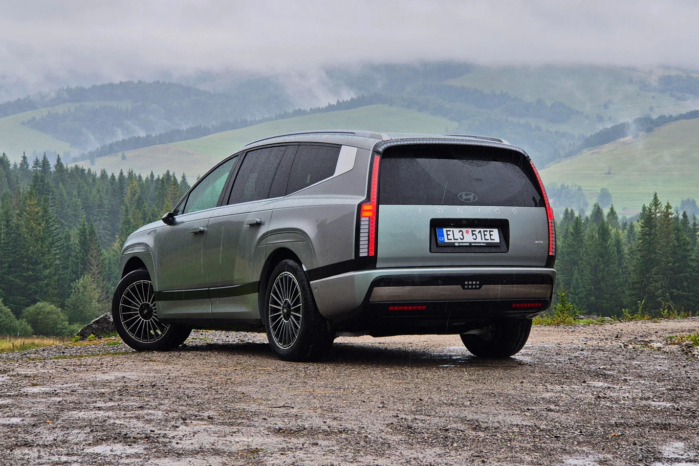 Nowoczesny, srebrny SUV Hyundai stoi na szutrowej drodze z widokiem na las i górzysty krajobraz w tle, pojazd posiada czarne felgi i charakterystyczne tylne światła LED.