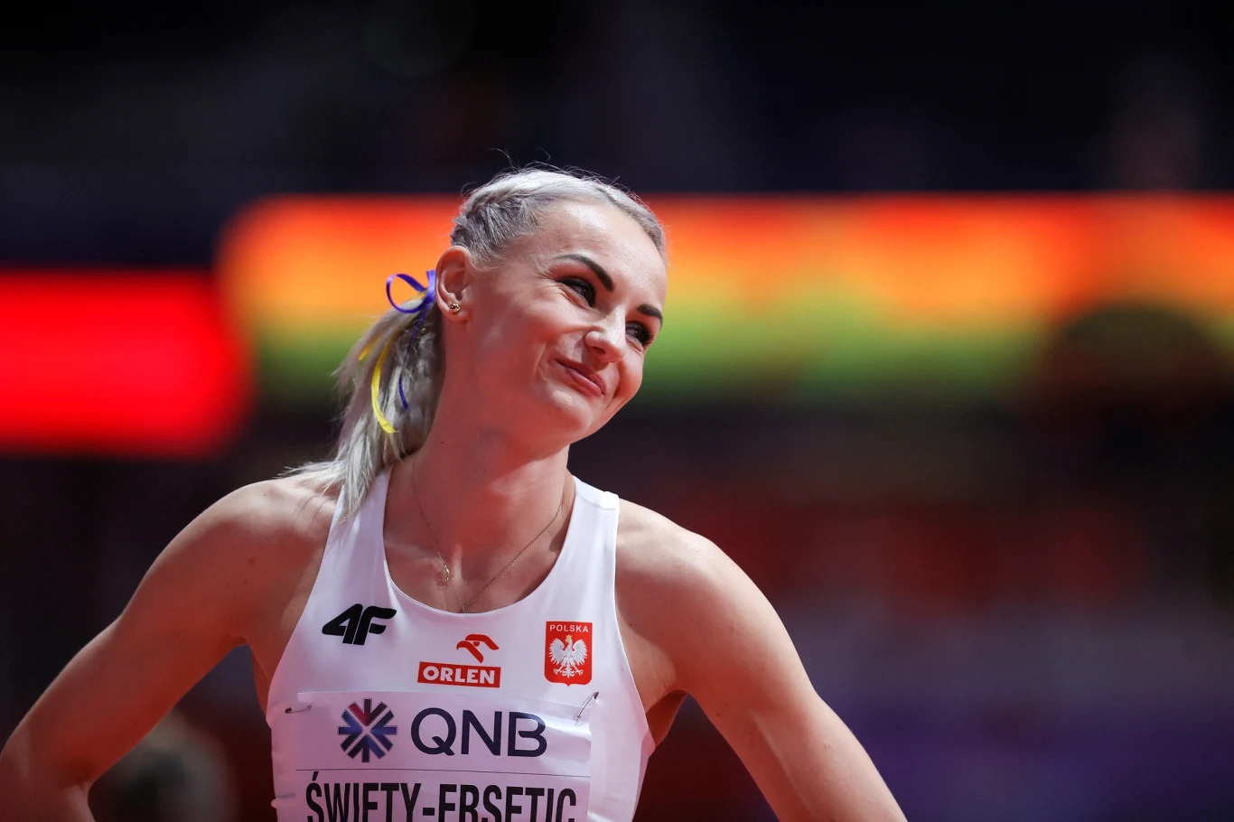 Uśmiechnięta sportsmenka w białym stroju reprezentacji Polski na arenie sportowej, z widocznym napisem Polska oraz numerem startowym, w tle rozmyte barwy trybun.