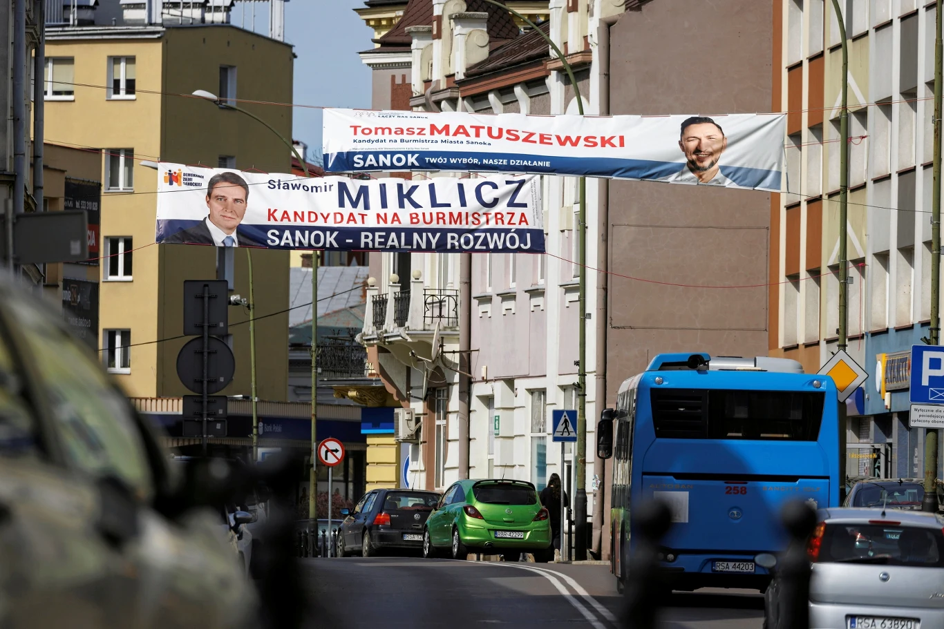 Tomasz Matuszewski i Sławomir Miklicz stanęli w szranki o fotel burmistrza Sanoka. Zwycięzcą z 50,73 proc. głosów na koncie w II turze okazał się pierwszy z nich Ulica miejska z zawieszonymi banerami wyborczymi dwóch kandydatów na burmistrza, samochody na jezdni, piesi na chodniku, w tle widoczne budynki mieszkalne oraz autobus.