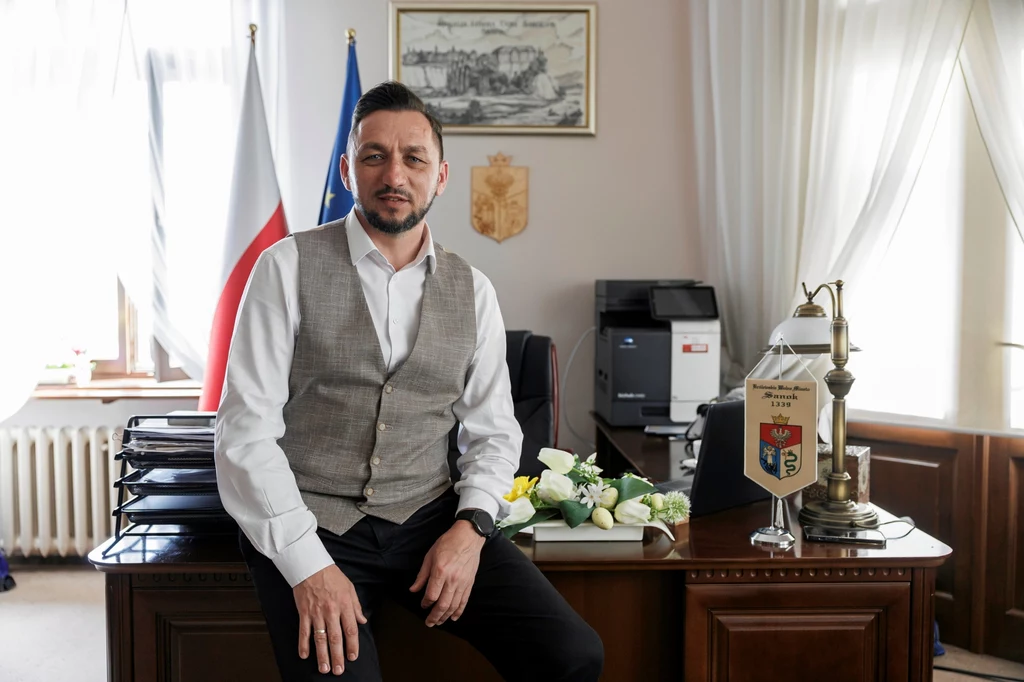 Mężczyzna w koszuli i kamizelce siedzi na biurku w eleganckim gabinecie urzędowym, w tle polska i unijna flaga, oficjalne dokumenty oraz dekoracje biurowe, podkreślające charakter miejsca pracy.