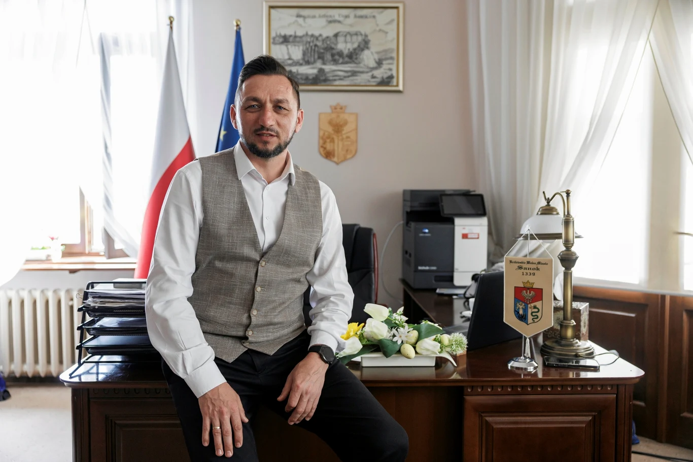 Burmistrz Sanoka Tomasz Matuszewski Mężczyzna w koszuli i kamizelce siedzi na biurku w eleganckim gabinecie urzędowym, w tle polska i unijna flaga, oficjalne dokumenty oraz dekoracje biurowe, podkreślające charakter miejsca pracy.