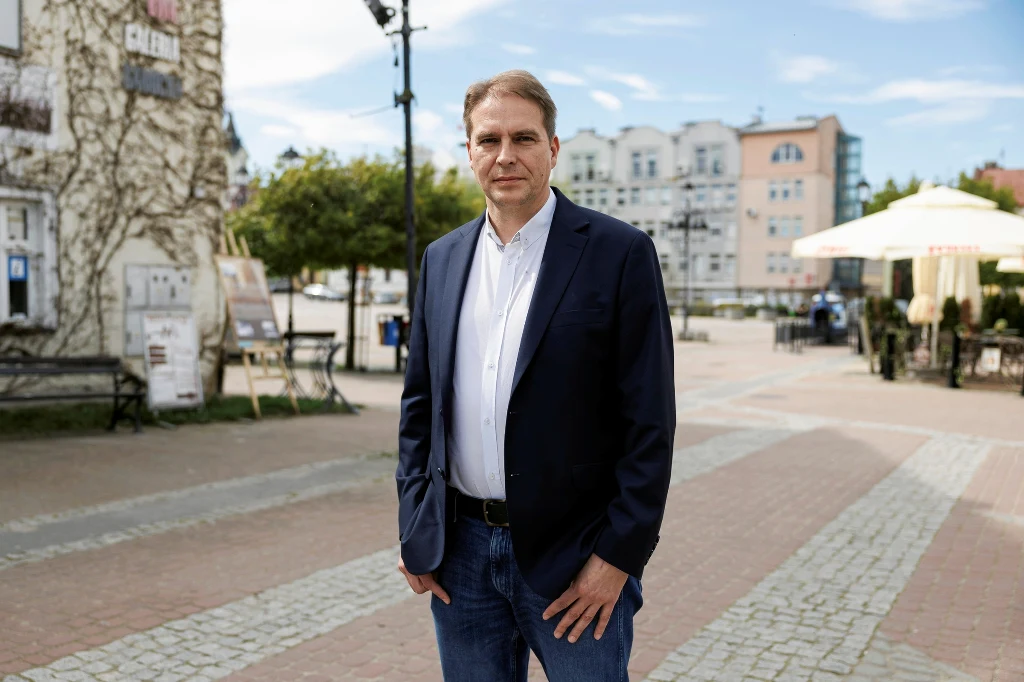 Mężczyzna w średnim wieku w granatowej marynarce i białej koszuli stoi na pustym rynku miejskim z zabudową mieszkaniową w tle oraz kawiarnią z parasolami po prawej stronie.