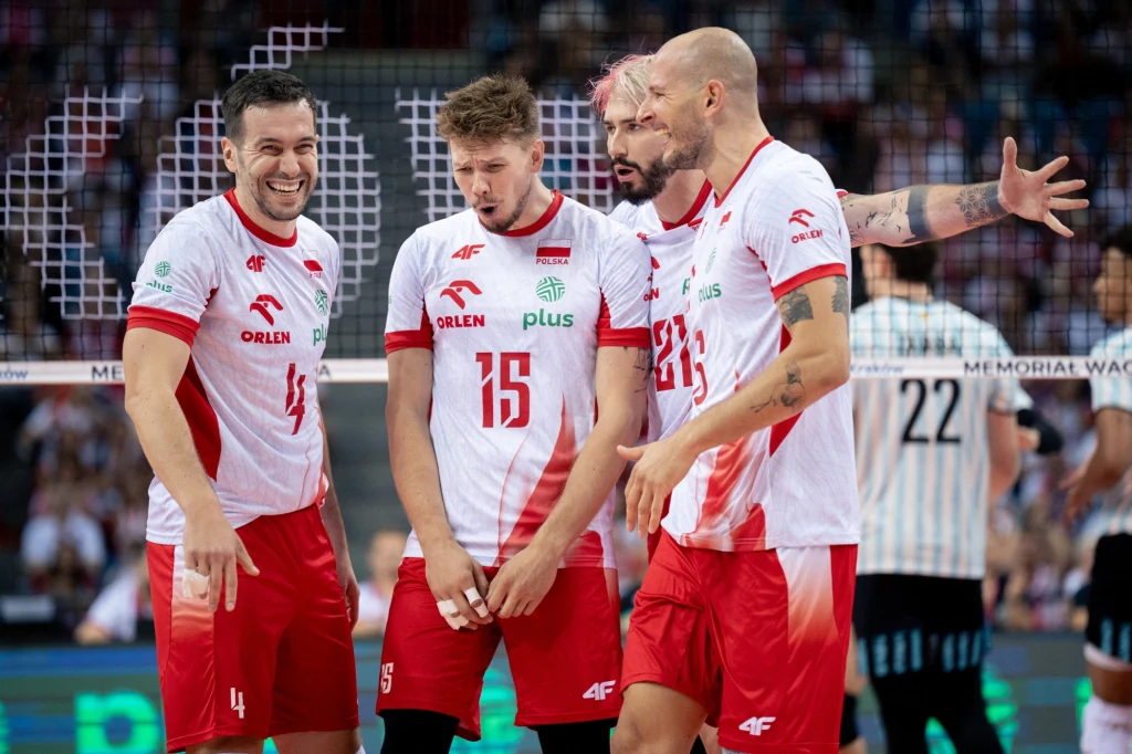 Grupa polskich siatkarzy w białych koszulkach z czerwonymi elementami świętuje zdobycie punktu podczas meczu, zawodnicy okazują emocje, w tle widoczna siatka oraz sylwetki przeciwników.