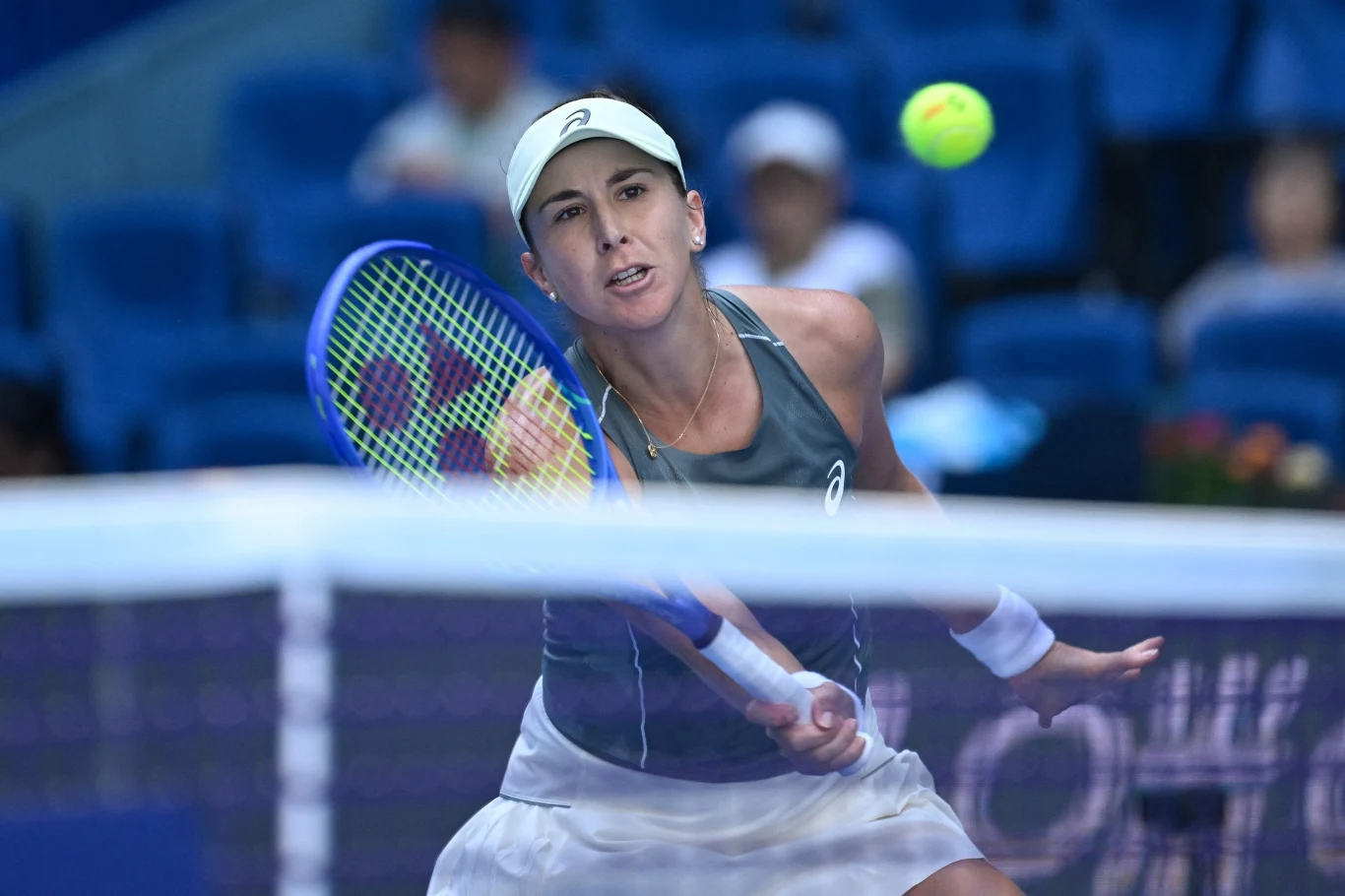 Tenisistka uderzająca piłkę rakietą podczas meczu na korcie, skoncentrowana na akcji, w tle widoczne rozmyte postacie widzów.