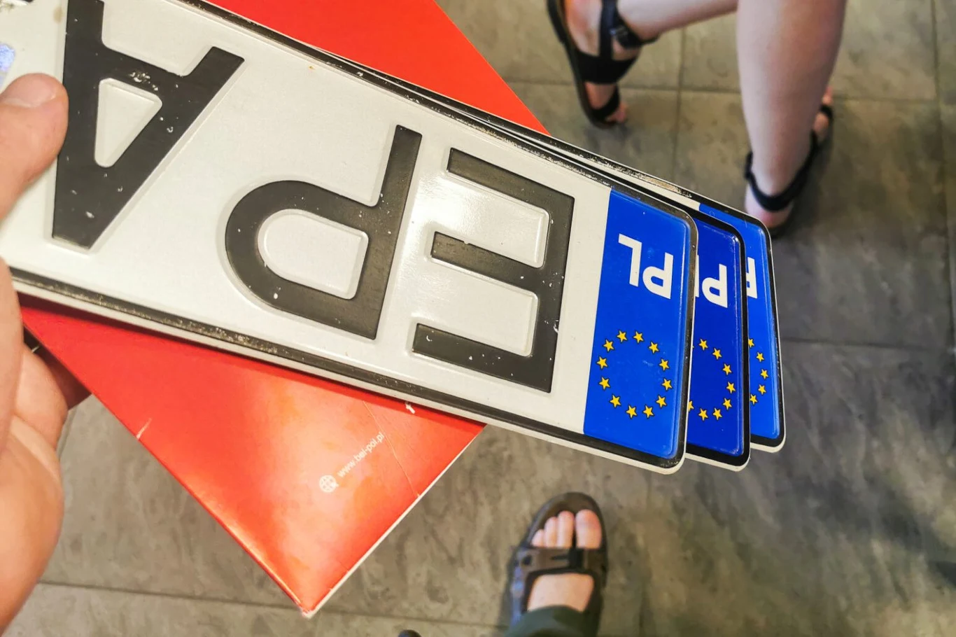 Wyższe stawki za wydanie dokumentów i tablic rejestracyjnych. Kierowcy sięgną do kieszeni. Ręka trzymająca trzy polskie tablice rejestracyjne z oznaczeniem „PL” i flagą Unii Europejskiej, w tle fragmenty nóg osób w sandałach oraz podłoga o ciemnym wzorze.