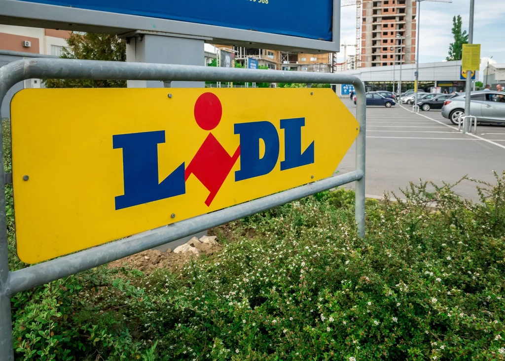 Żółta tablica z logo i nazwą Lidla w formie wskaźnika kierunku, otoczona zielonymi krzewami, w tle parking i budynki wielorodzinne.