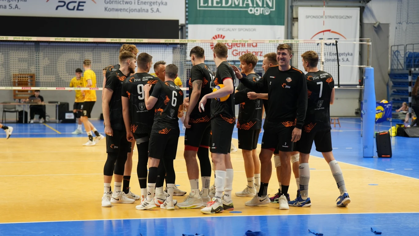 Grupa siatkarzy w czarnych strojach drużynowych zebrała się na boisku podczas meczu halowego, widoczne emocje i zgranie zespołu, w tle napisy sponsorów oraz fragment siatki.